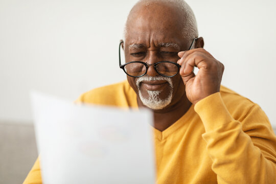 An elderly man wearing a yellow sweater focuses intently as he reads a document. He adjusts his glasses, looking thoughtful and engaged in a comfortable indoor space.