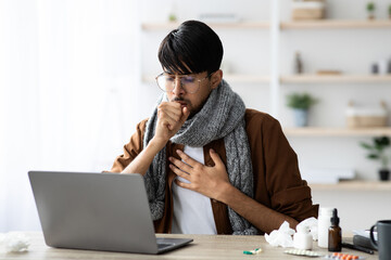 A young man wearing a scarf is seated at a wooden desk, coughing while focusing on a laptop screen in a bright indoor space. Tissue paper and medicine are visible.