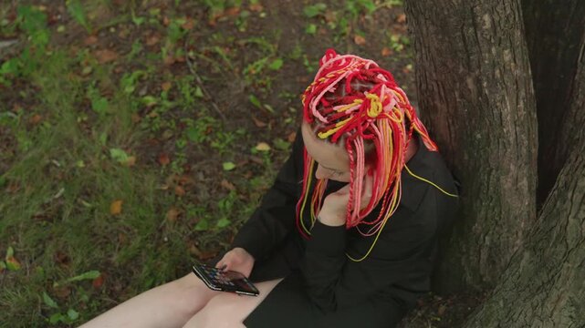 wide-shot caucasian woman with neon dreadlocks sitting under tree, looking at phone with headphones, park leaves around legs, muted coat contrasts vibrant hair, scrolling gesture conveys isolation