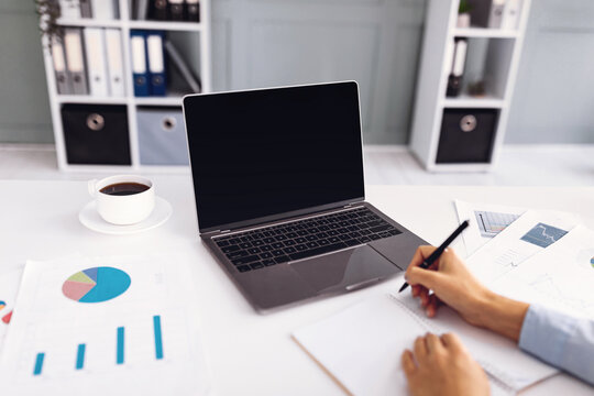 A person writes notes and analyzes graphs at a clean office desk. A laptop sits open next to coffee, with various charts and documents displayed nearby. - Powered by Adobe