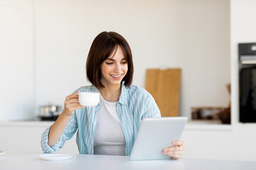 Bright morning light fills a modern kitchen as a woman smiles while holding a cup of coffee and looking at a tablet. She appears relaxed and engaged with her device.