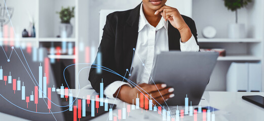 In a modern office, a confident black woman dressed in formal attire engages with a digital tablet. She focuses on business planning and productivity while surrounded by stylish decor.