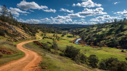 Fototapeta premium The scene shows a winding dirt path beside a river in a valley. Lush green hills rise on either side with blue sky and scattered clouds overhead.
