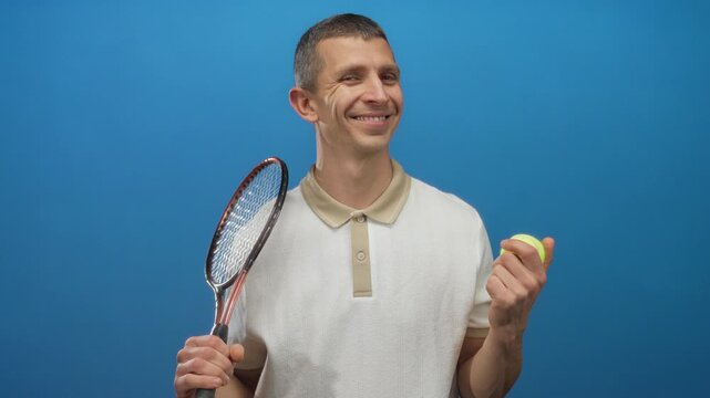 Caucasian man holding tennis racket and ball against blue background, expressing positive emotion in isolated wall setting, highlighting sport and athleticism.