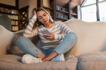 Obraz premium A young woman is seated on a soft couch, her legs crossed, as she gazes intently at her smartphone, appearing deep in thought. The living room is warm and inviting, with natural light coming in.