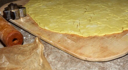 Cookie dough rolled flat on a wooden board, cut into Christmas tree shapes. A metal cutter and rolling pin lie nearby, showing the process of preparing holiday cookies at home.