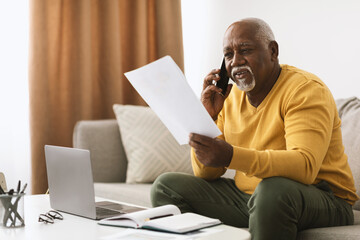 A senior man sits on a sofa, speaking on the phone while examining papers in his hand. A laptop and notebook are on the table in front of him, indicating a busy day at home.