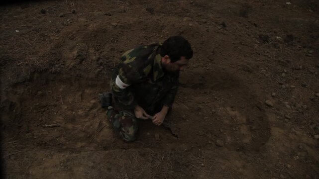 An exhausted soldier in camouflage buries a fallen comrade in a forest trench. He frantically covers the body with dirt using a shovel, surrounded by silent tall pine trees.

