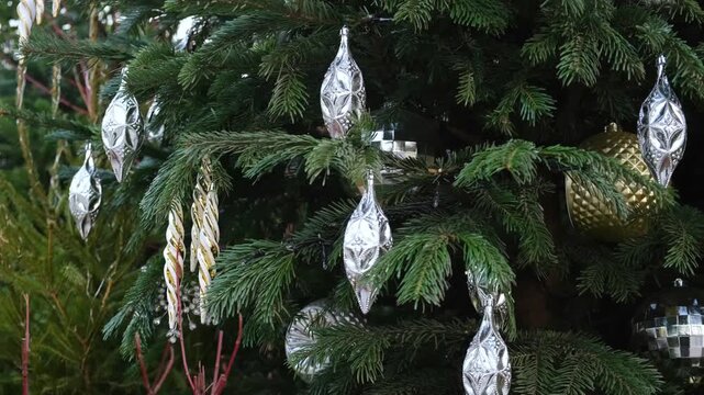Silver vintage ornaments and twisted icicles gently swaying on evergreen branches in the wind, elegant outdoor Christmas tree decoration for city holiday mood, shopping street decor, and winter celebr