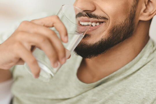 A man smiles as he sips from a clear glass of water, resting comfortably in a cozy indoor space. The setting suggests a moment of relaxation and refreshment.
