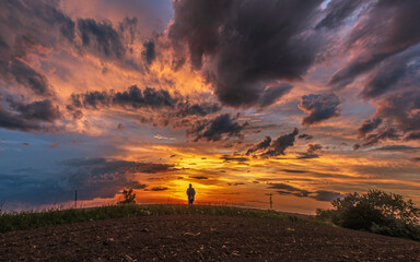 Man at Sunset Landscape – A lone man stands before a dramatic sunset sky, framed by vibrant clouds over an open field, symbolizing the connection between humans and nature.