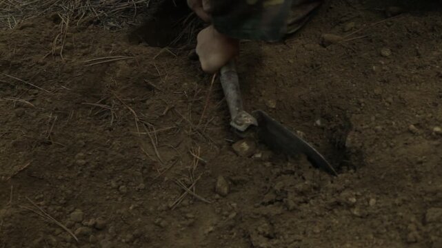 An exhausted soldier digs a grave in a pine forest. Overcome with emotion and physical fatigue, he collapses onto the fresh dirt, struggling to continue his grim task.


