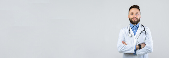 A male doctor with a beard and a stethoscope around his neck is smiling and standing confidently. He has arms crossed in a neutral space, wearing a white lab coat and blue shirt.