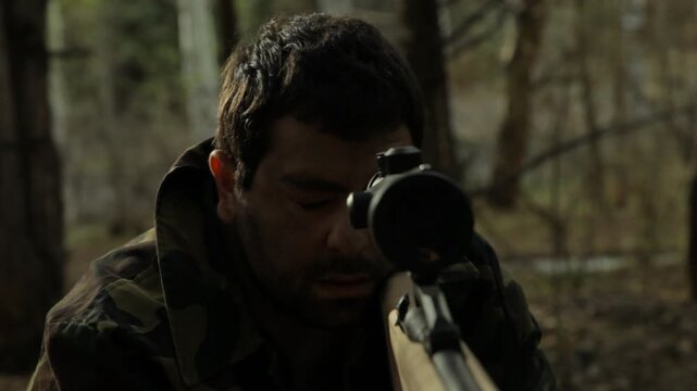  Close-up of hunter looking through rifle scope in forest. He wears camouflage jacket. He holds rifle firmly and peers intently down barrel. Fall day.