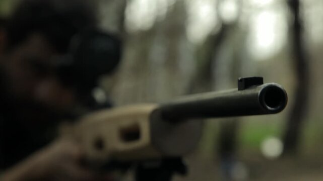  Close-up of hunter looking through rifle scope in forest. He wears camouflage jacket. He holds rifle firmly and peers intently down barrel. Fall day.
