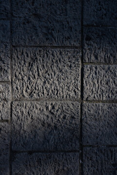 A dark, moody photograph of chiseled stone tiles arranged in a grid-like structure. Subtle directional light highlights the carved texture and variations in each slab, emphasizing craftsmanship. 