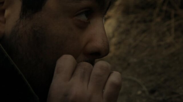 Close-up of nervous bearded soldier hiding in earthen trench. He smokes cigarette with shaking hands, eyes darting fearfully. He glances at silent walkie-talkie on the ground.