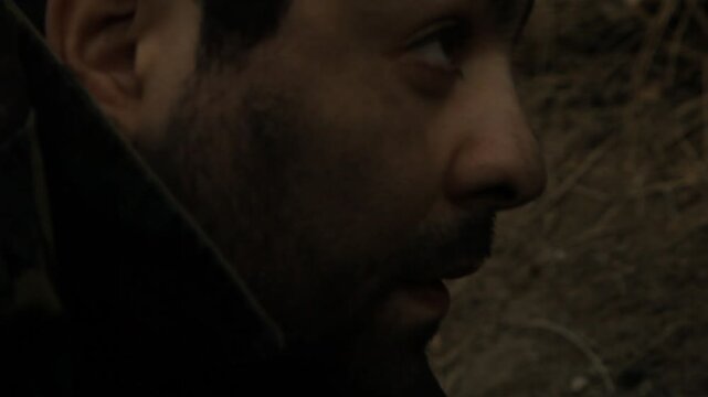 Close-up of nervous bearded soldier hiding in earthen trench. He smokes cigarette with shaking hands, eyes darting fearfully. He glances at silent walkie-talkie on the ground.
