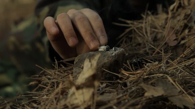 Close-up of camouflaged soldier placing lit cigarette on rock next to rifle barrel. He leaves it smoking on the stone to grip his weapon with both hands.