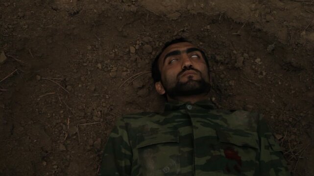 Top view of a dead soldier falling into a grave. The man in camouflage uniform has been shot and lies lifeless with wide open eyes on the dirt.