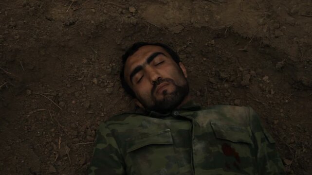 Top view of a dead soldier falling into a grave. The man in camouflage uniform has been shot and lies lifeless with wide open eyes on the dirt.