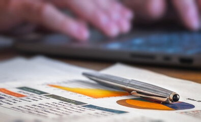 Pen on financial charts with laptop background