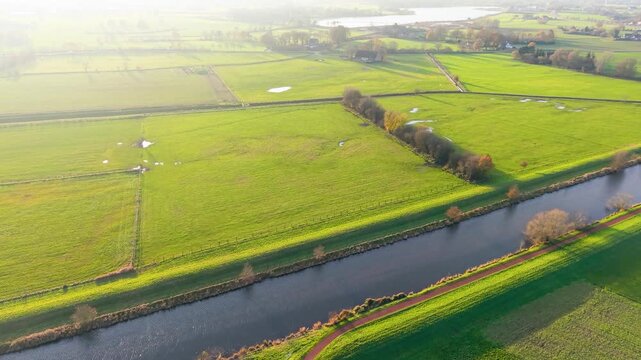 Aerial view of rural fields divided by hedgerows, canal, and scattered trees sunlit landscape with water patches, paths, and distant farmhouses in vibrant countryside.