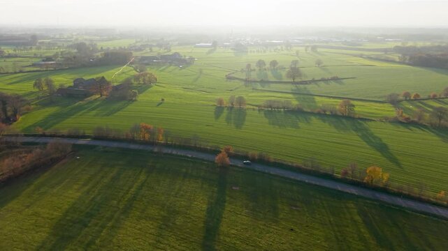 Aerial view of sunlit rural fields with winding road, scattered trees, and long shadows tranquil countryside scene with soft haze and warm morning light.