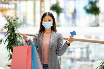 A woman stands in a bright mall holding shopping bags in one hand and a credit card in the other. She is wearing a mask, and the atmosphere is lively with shoppers around.