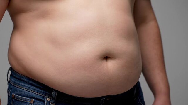 Close up of a man's overweight abdomen showing belly fat and blue denim jeans against a plain grey studio background symbolizing health concerns and body image issues with soft studio lighting.