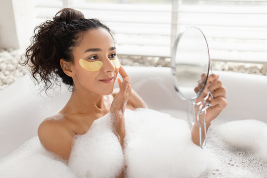 A woman enjoying a soothing bubble bath in a stylish bathroom, applying skincare patches while looking into a mirror, enhancing her self-care routine. - Powered by Adobe