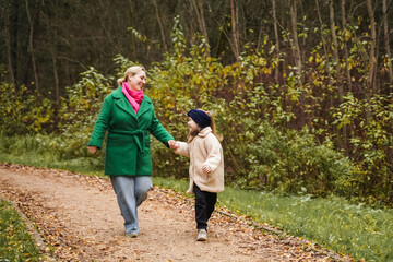 Fototapeta premium Walking mother with daughter on park path. Holding hands shows parenting, care, trust, safety. Outdoor family activity supports bonding, happiness, healthy lifestyle, weekend leisure, forest trail.