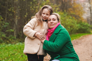 Fototapeta premium Cuddling mother with daughter in park. Smiling family portrait shows love, affection, support. Close bonding supports childhood happiness, motherhood care, trust, safe relationship, outdoor lifestyle.
