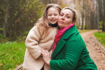 Fototapeta premium Cuddling mother with daughter in park. Smiling family portrait shows love, affection, support. Close bonding supports childhood happiness, motherhood care, trust, safe relationship, outdoor lifestyle.