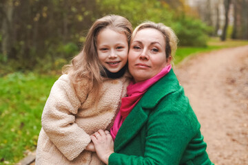 Fototapeta premium Cuddling mother with daughter in park. Smiling family portrait shows love, affection, support. Close bonding supports childhood happiness, motherhood care, trust, safe relationship, outdoor lifestyle.