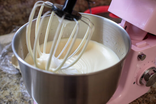 Close-up of a whisk attachment in a pink stand mixer efficiently whipping a bowl of white cream or batter to a fluffy consistency in a stainless steel bowl