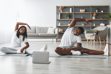 Young black couple warming up stretching arms watching fitness video tutorial online on laptop, guy and lady doing workout sitting on floor mat in living room practicing yoga side bending exercise