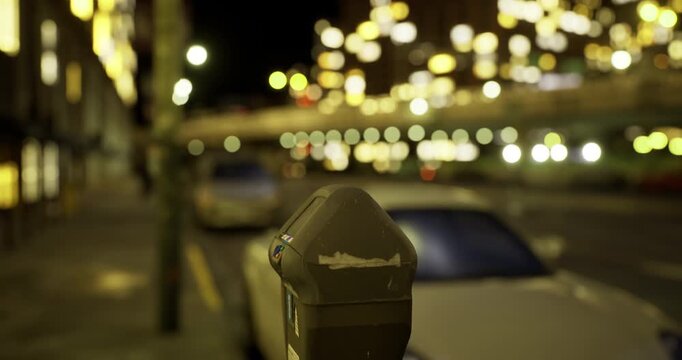 Streetlights illuminate a quiet city road, creating a dreamy atmosphere at night. A parking meter stands in focus, while blurred vehicles and lights enhance the tranquil vibe of the scene.