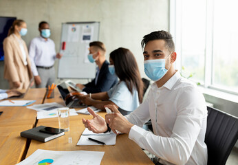 Arab businessman wearing a medical mask uses disinfectant spray while participating in a meeting. Colleagues also follow safety protocols in a modern boardroom setting.