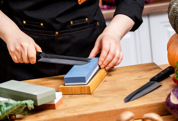 Focused woman in black chef apron sharpens knife on blue whetstone at wooden kitchen table with colorful vegetables. Cooking, culinary technique, healthy lifestyle concept.