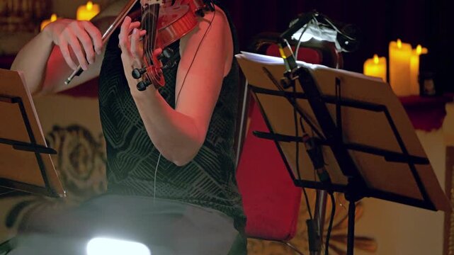 A front view of a woman in a dark dress playing the violin. Candles burn in the background. The musician's hands move elegantly. A classical music concert