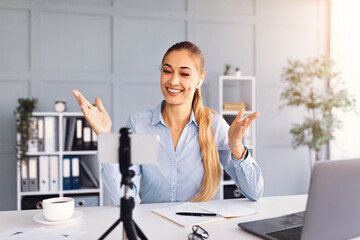 A woman smiles and gestures while participating in a video call from her home office. A laptop, notebook, and coffee cup are visible on her desk, creating a comfortable workspace.