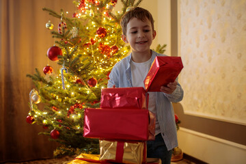 Happy young boy in a striped shirt standing proudly next to a tall stack of red and gold Christmas...