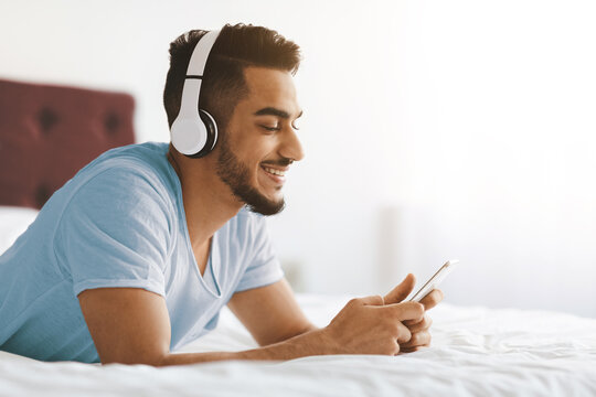 A young man lies on his stomach on a bed, wearing headphones and smiling as he listens to music on his smartphone. The bright room creates a cheerful atmosphere. - Powered by Adobe