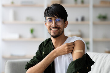 A young man sits comfortably in a bright living room, smiling while pulling up his sleeve to show a small bandage on his arm after receiving a vaccination.