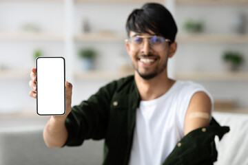 A smiling young man with glasses displays his smartphone proudly in a bright, comfortable room. A vaccination bandage is visible on his arm, suggesting recent immunization.