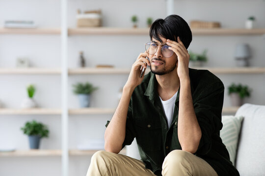 A young man wearing glasses sits on a couch, holding a phone to his ear and rubbing his forehead, showing signs of stress during a conversation in a stylish room. - Powered by Adobe