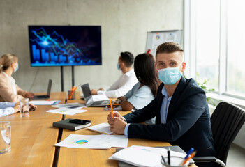 Diverse group of coworkers wearing medical masks engaged in a conference in an office. They discuss charts and data while navigating challenges during the health pandemic.