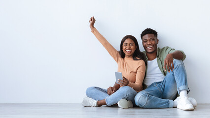 Two happy individuals sit together on a smooth floor, one holding a smartphone and smiling widely. Their expressions convey joy and excitement in a bright, airy room.