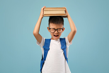Obraz premium Young boy wearing glasses and a backpack holds a stack of books above his head with a frustrated expression, illustrating the challenge of carrying school materials.